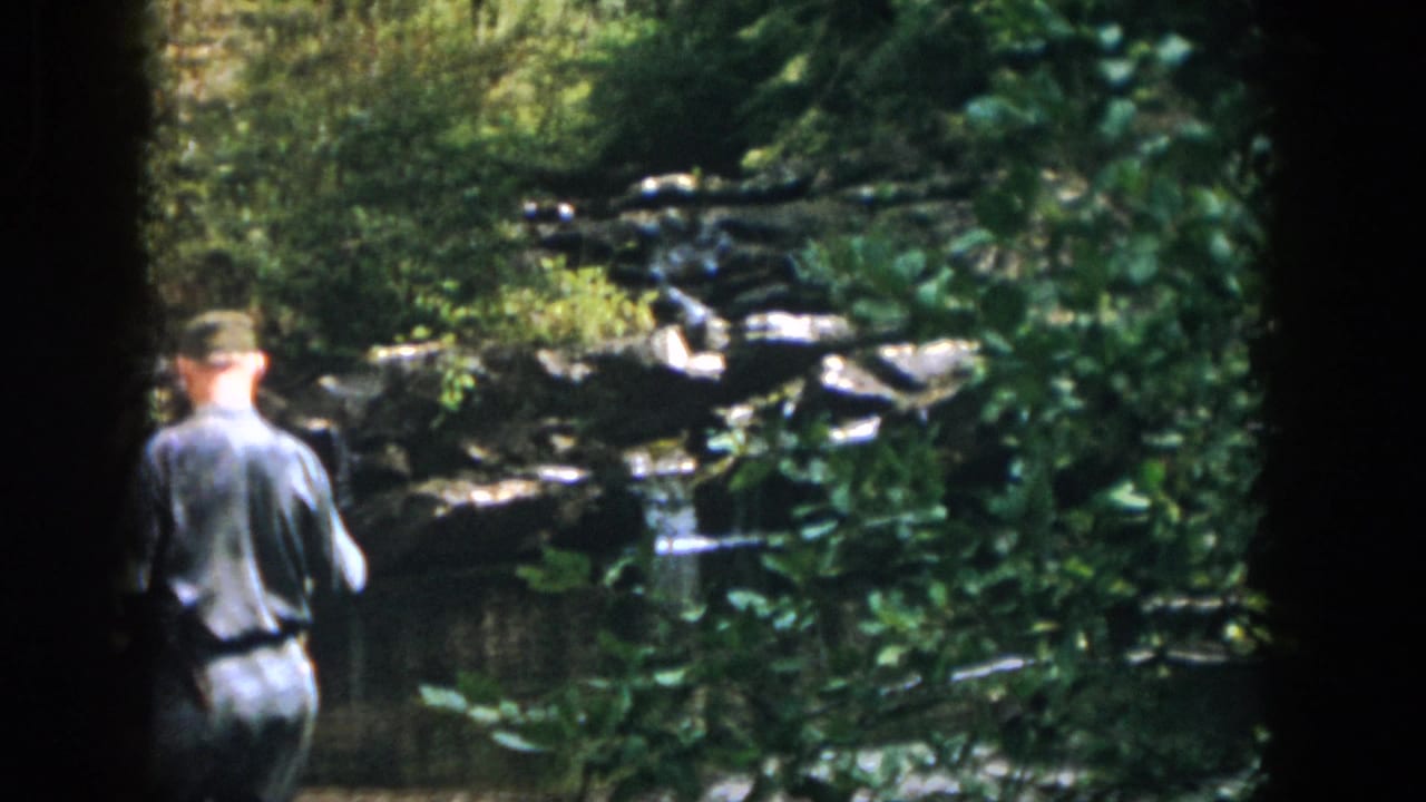A uniformed man in 1958 Dayton, Ohio footage stands near a rocky pond surrounded by trees and brush.