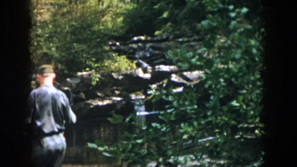 A uniformed man in 1958 Dayton, Ohio footage stands near a rocky pond surrounded by trees and brush.