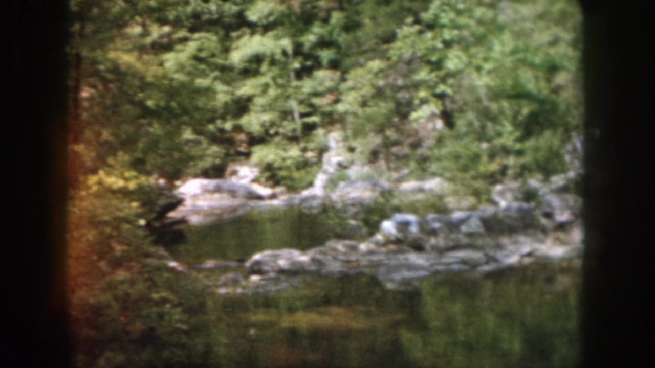 1958 Dayton-area home-movie frame showing a quiet rocky river pool bordered by dense green trees and foliage.