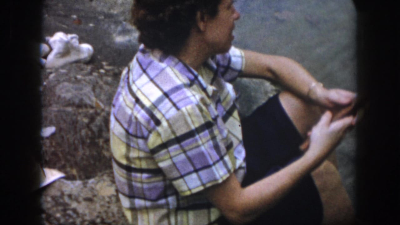 Adult in a plaid shirt and dark shorts seated beside shallow water on rocks.