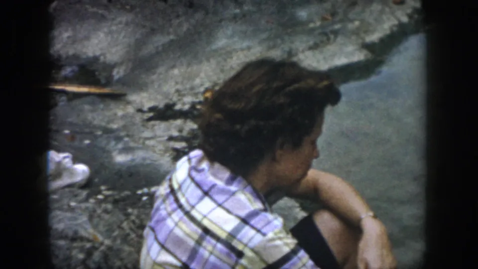 The seated adult leans toward the water on a rocky bank, with a fishing rod visible along the edge of the frame.