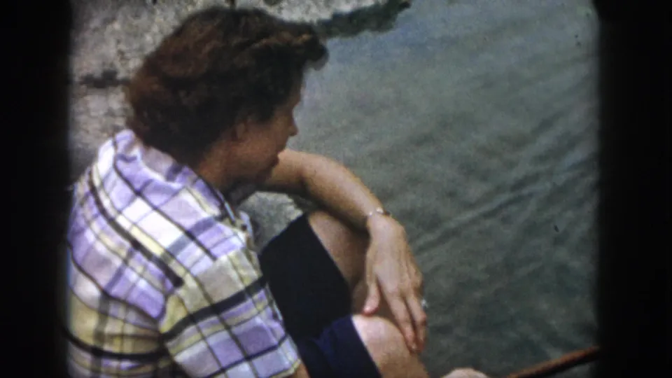 A 1958 Dayton home-movie frame shows the adult seated beside rippled river water, arms resting casually on bent knees.