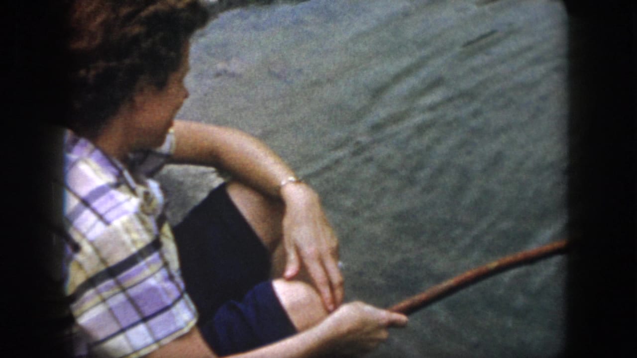 In 1958 Dayton, Ohio home-movie footage, a seated person in a plaid top rests by pond water with a stick in hand.