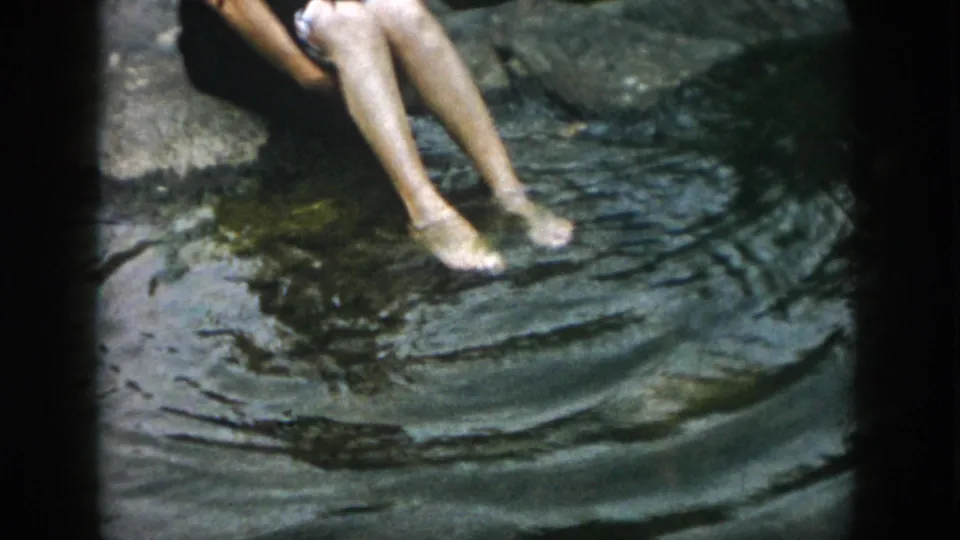 The seated person rests barefoot at the pond edge, with legs and feet visible in the water in this 1958 Dayton home movie.