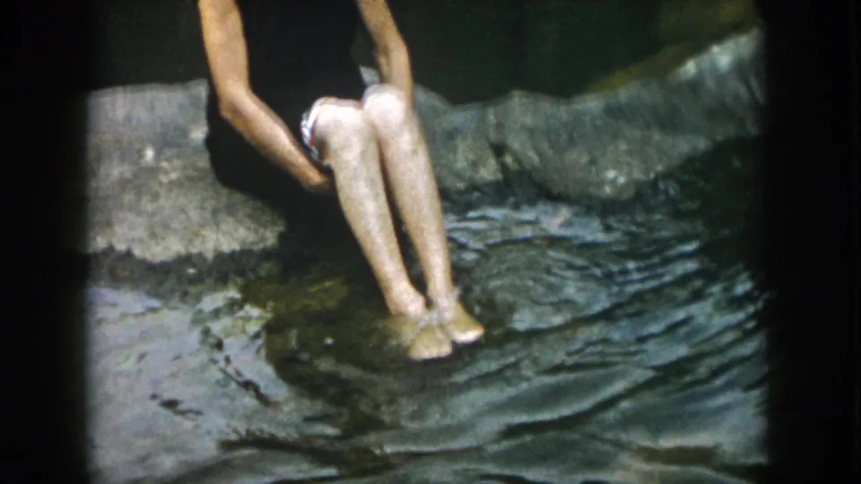 A wider frame shows the person seated beside the pond, bare feet in the rippling water, in the 1958 Dayton, Ohio footage.