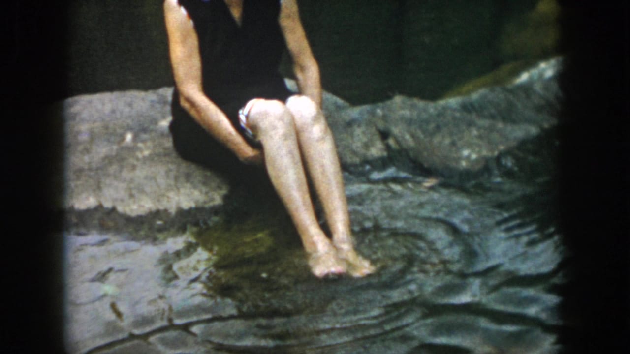 1958 Dayton-area 8mm frame showing a woman in a dark sleeveless swimsuit sitting on a rock beside a shallow stream.
