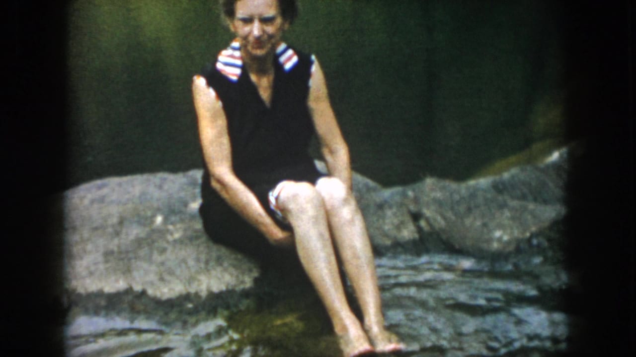 A woman in a dark sleeveless swimsuit with a striped collar sits barefoot on rocks by shallow water in 1958 home-movie footage.
