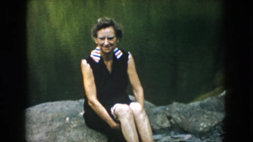 1958 Dayton, Ohio home-movie frame of a woman seated on a large rock with a shallow stream and green background behind her.