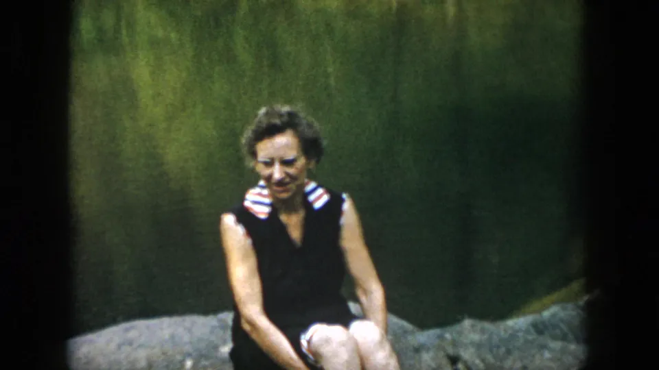 A soft 8mm frame shows the woman in a dark swimsuit sitting beside water, with rocks and a green natural backdrop visible.