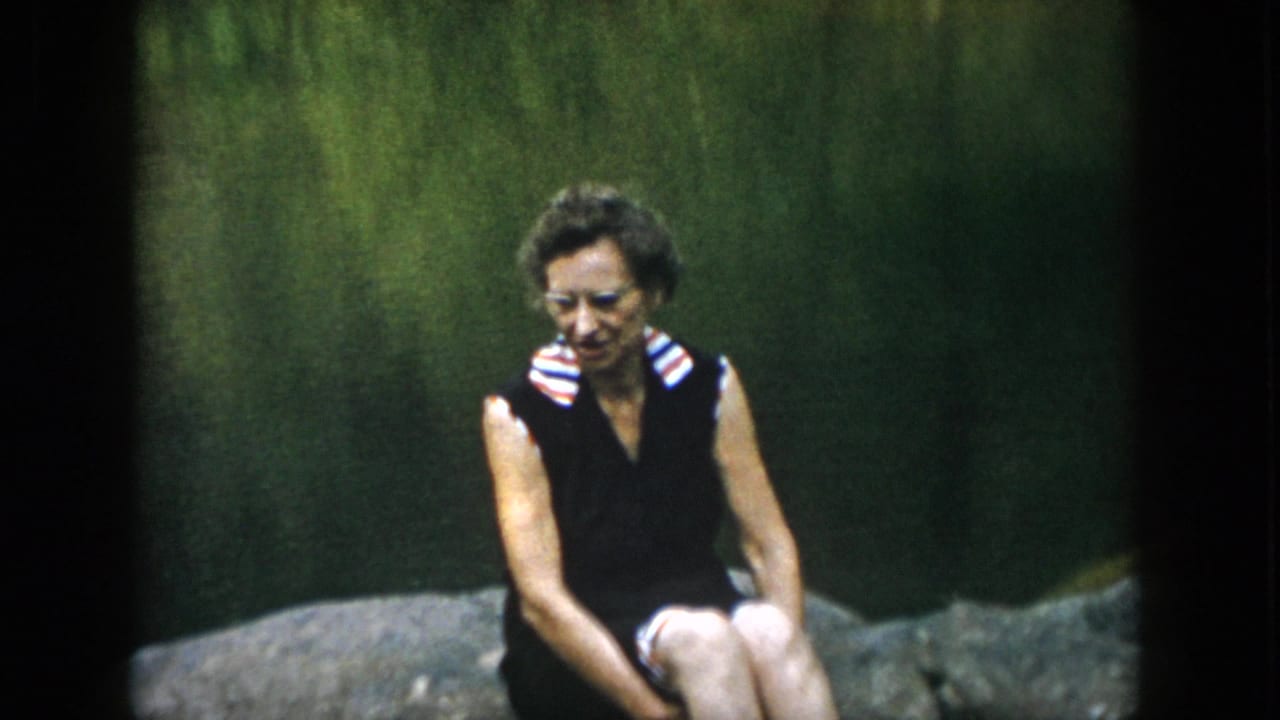 In 1958 Dayton home-movie footage, a woman wearing glasses and a dark sleeveless dress sits on a large rock near a green lakeside backdrop.