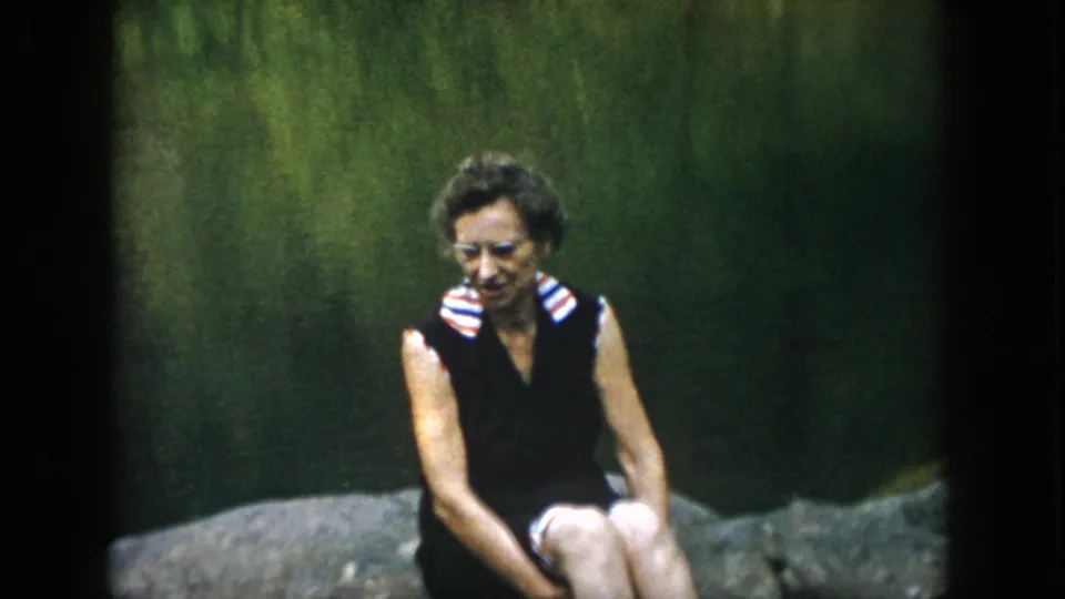 In 1958 Dayton home-movie footage, a woman wearing glasses and a dark sleeveless dress sits on a large rock near a green lakeside backdrop.