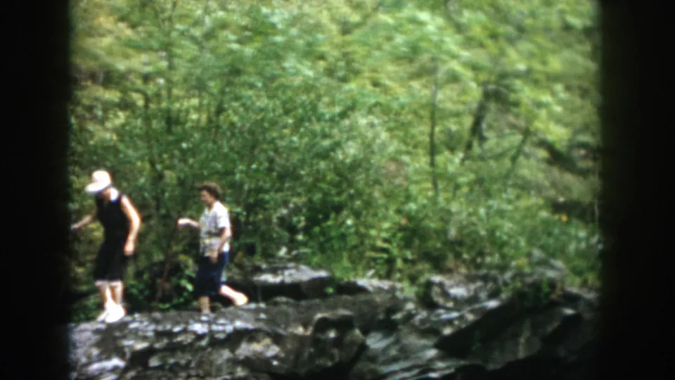 Two adults move carefully over rocks in a wooded area, with dense green foliage and a stream or rocky shoreline visible in the 1958 home movie.