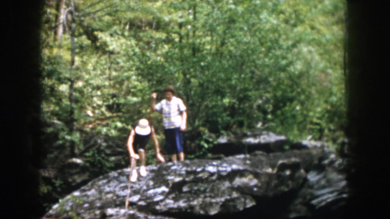 A hiker in a white hat bends near rocks while another stands nearby.