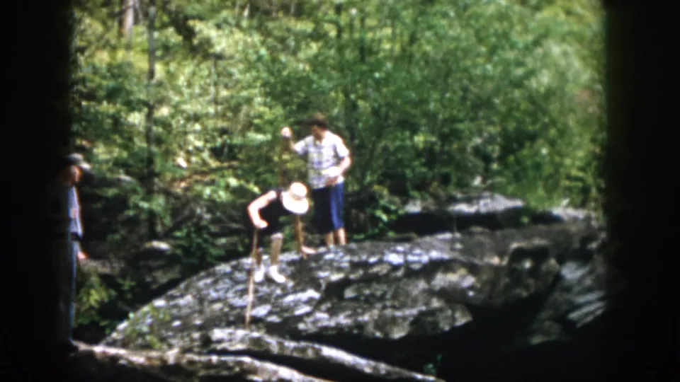 1958 Dayton, Ohio home-movie frame of adults navigating a rocky forest trail, with a third partial figure at the edge.