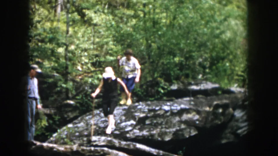 1958 Dayton, Ohio home-movie frame showing a white-hatted adult stepping across rocky terrain while a companion is partly visible behind.