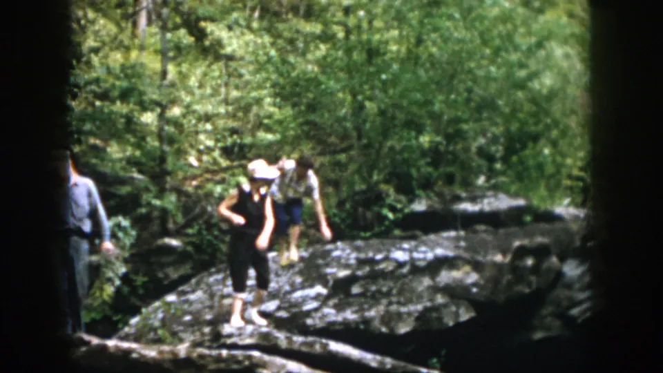 1958 home-movie frame from Dayton, Ohio showing two women moving carefully over large rocks in dense woods.