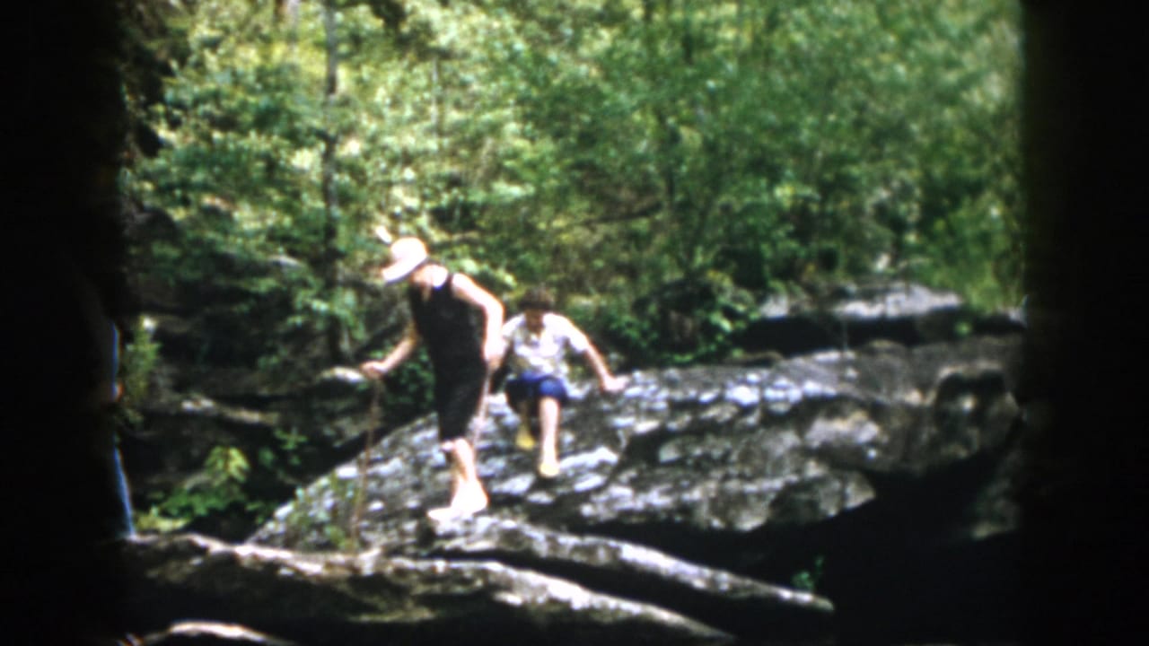 A woman in a white hat uses a stick while another woman follows on rocks.