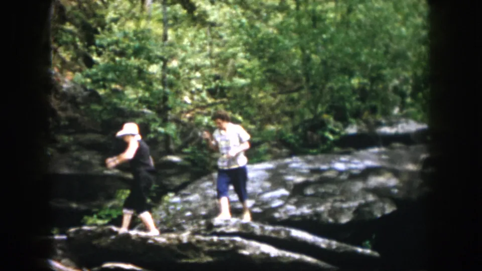 Home-movie frame of two women moving across a fallen branch and rocks, surrounded by leafy woods.