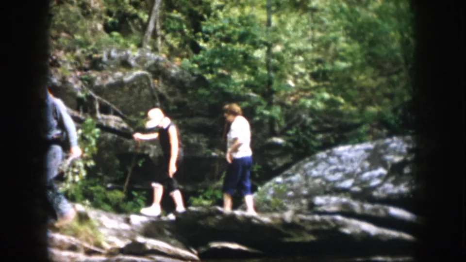 A 1958 Dayton, Ohio home-movie frame shows two women pausing on rocks in dense green woods near water.