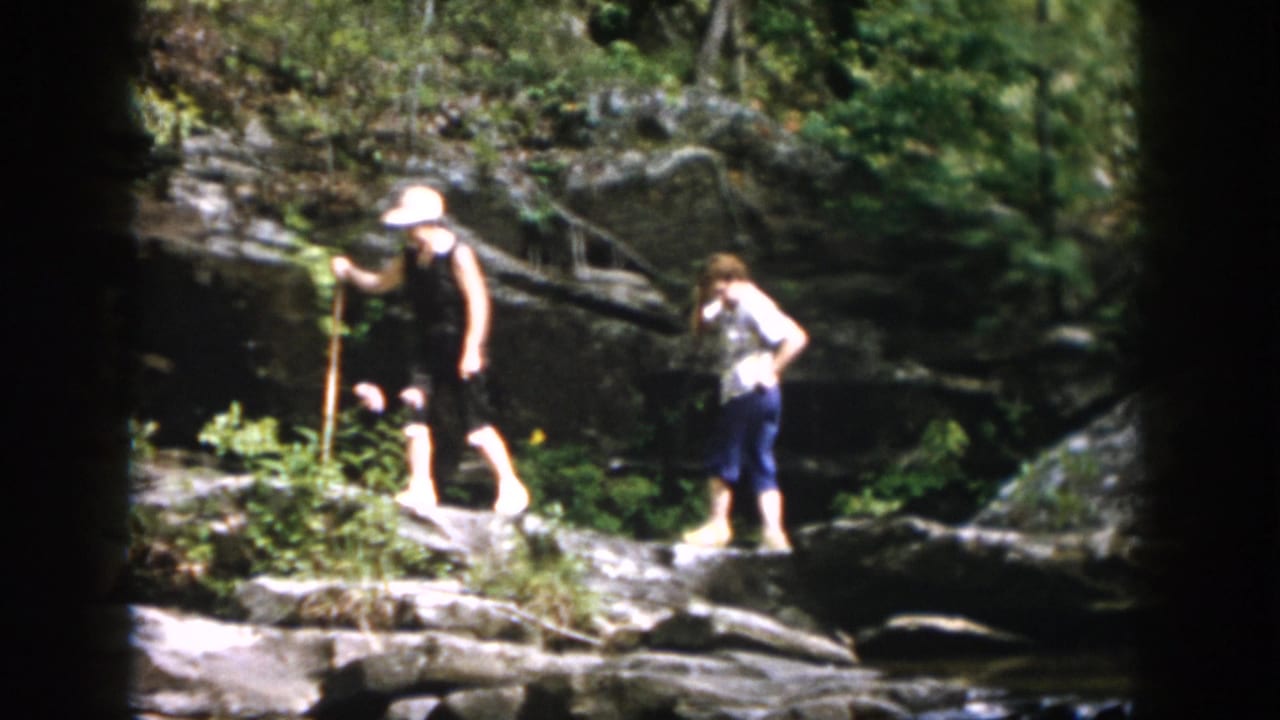 Two family members in a 1958 Dayton, Ohio home movie use walking sticks while crossing rocky ground by a forest stream.