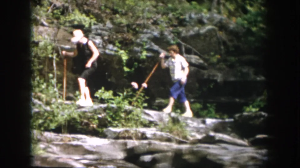 A mid-century home-movie frame shows two family members crossing rocks in a wooded stream setting near Dayton, Ohio.