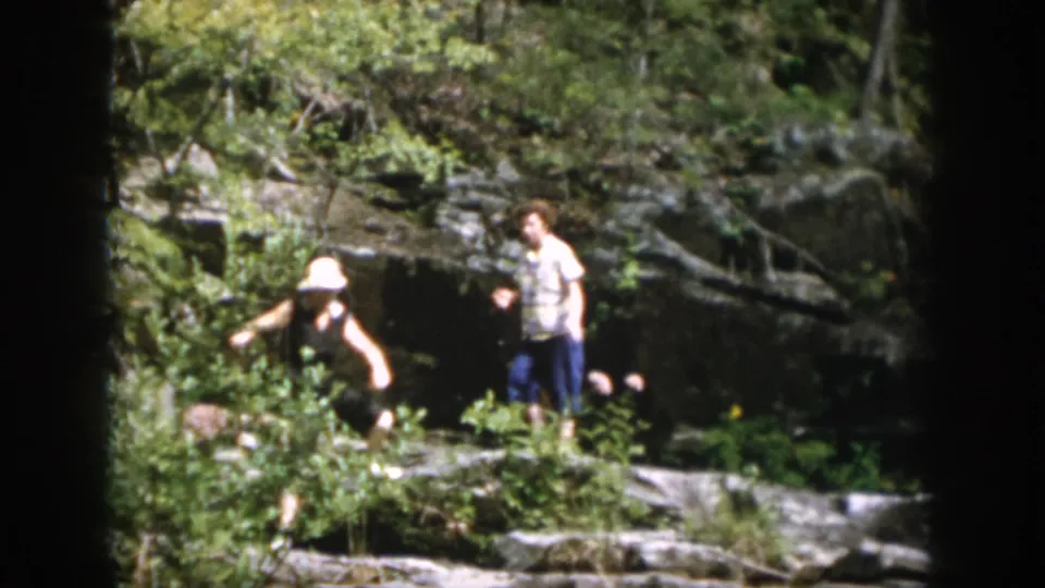The final frame shows a family member in a white hat stepping near brush and rocks while another person waits by the wooded stream.