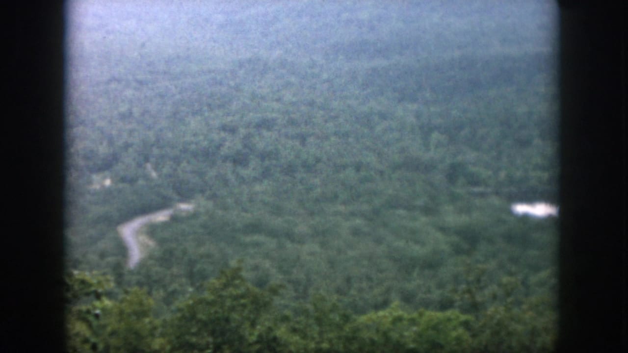 1958 8mm home-movie view across green hills, showing a curving road and a small bright water area beyond the trees.