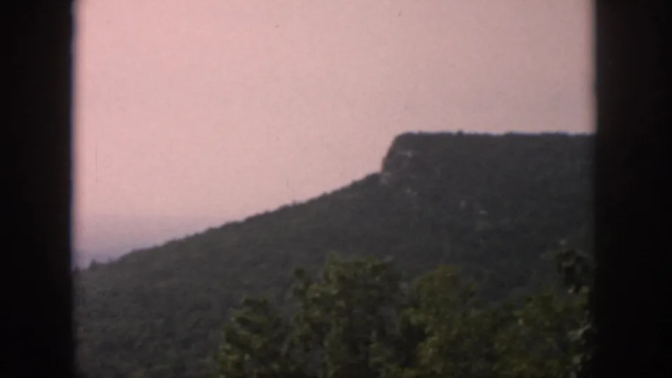 1958 8mm frame of a wooded ridge and distinct rocky outcrop, partially divided by dark window framing.