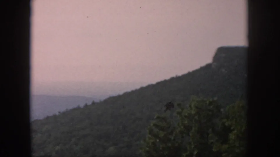 1958 8mm home-movie view of a dark forested ridge with a rocky top, shot through vertical window framing.
