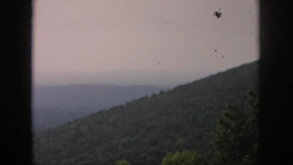 1958 8mm frame showing hazy distant hills beyond a wooded ridge, with dark window framing at the side.