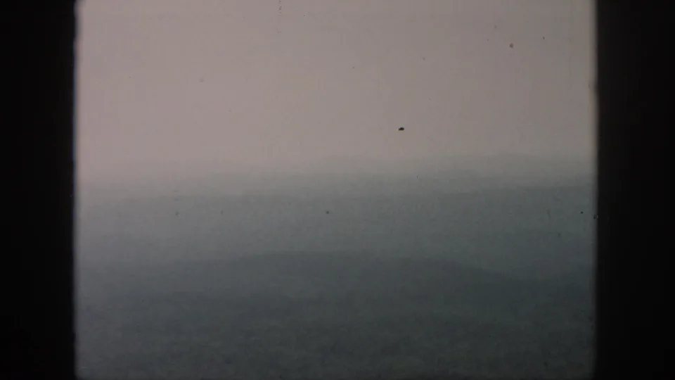 A foggy 8mm aerial view, attributed to Dayton, Ohio in 1958, with wooded hills softened by haze behind aircraft window bars.