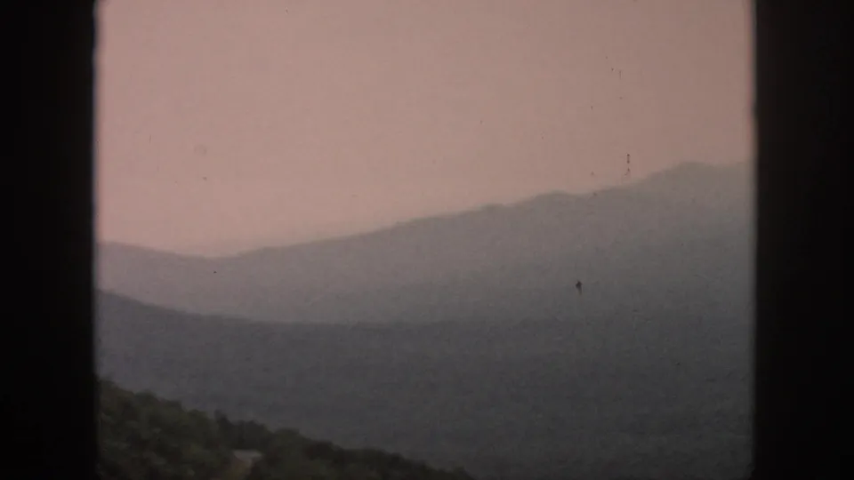 A 1958 home-movie view from an aircraft shows layered hills, forest, and a small pale road or clearing below.