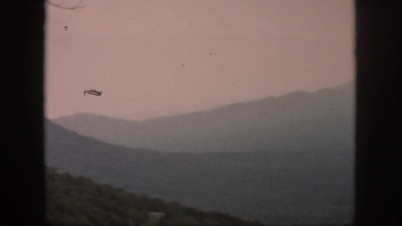 1958 8mm home-movie frame with hazy mountain ridges, wooded foreground hills, and dark window-like bars at the sides.