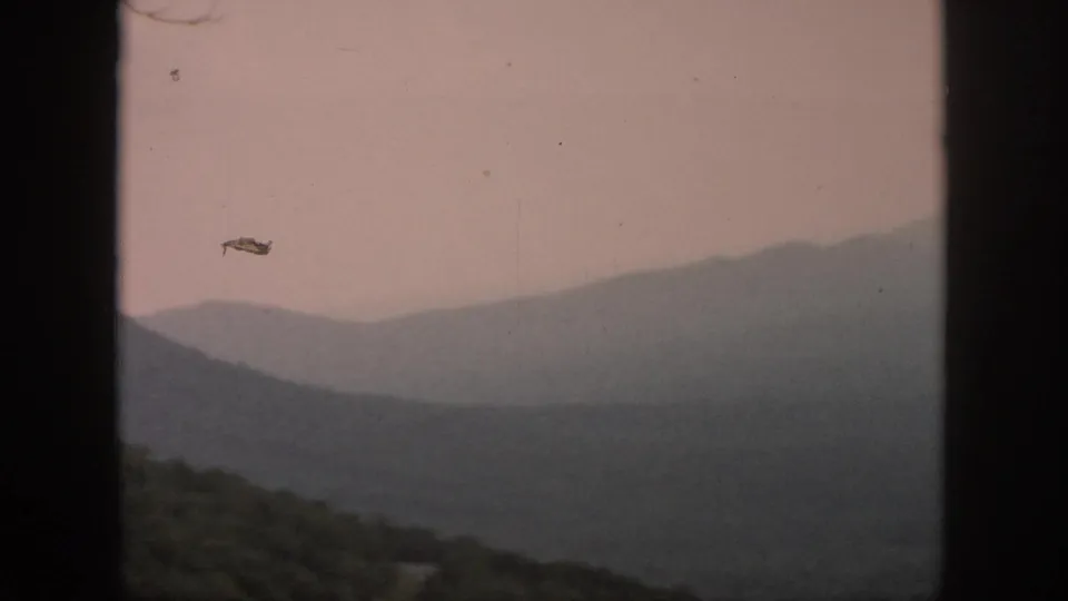 1958 8mm home-movie frame with hazy mountain ridges, wooded foreground hills, and dark window-like bars at the sides.
