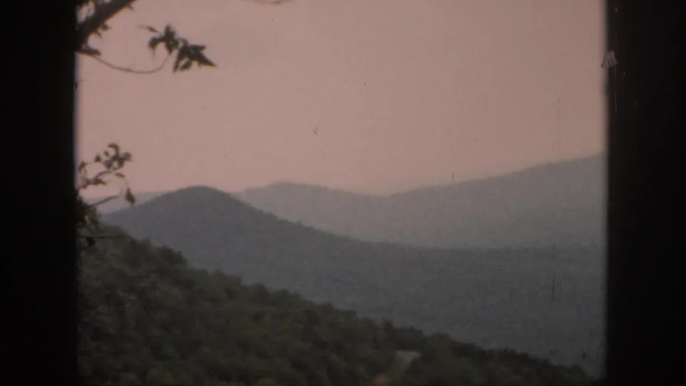 A 1958 8mm view of distant blue-gray ridges, a forested hillside, and small tree branches near the top edge.