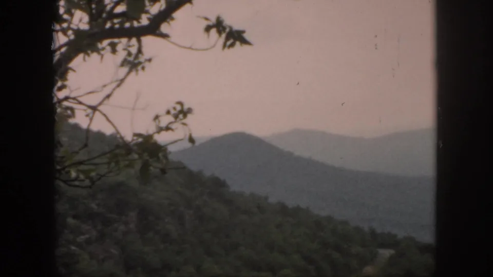 1958 home-movie frame showing a wooded hillside, foreground branches, and a rounded distant mountain ridge in soft haze.