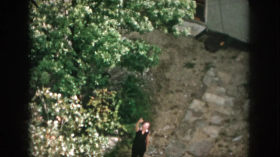 From above, a person waves near trees, rocks, and part of a modest building in the 1958 Dayton home-movie clip.