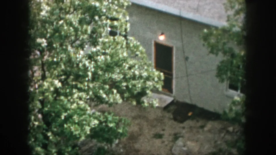 A modest house exterior with a lit light, open doorway or window, trees, and a rocky yard appears in the 1958 home-movie frame.