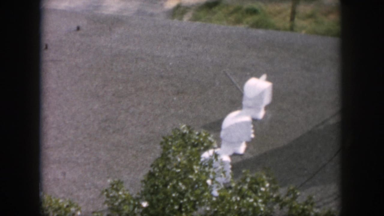1958 Dayton 8mm frame looking through a dark window toward white utility fixtures, pavement, and greenery outside.