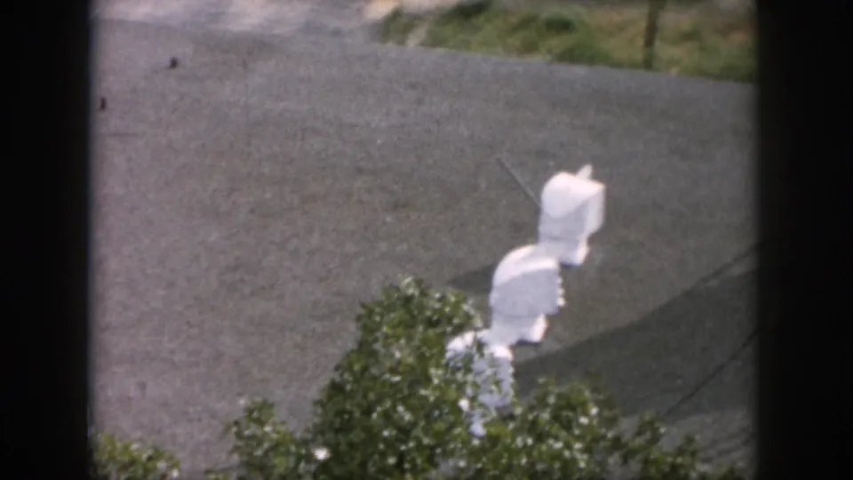 1958 Dayton 8mm frame looking through a dark window toward white utility fixtures, pavement, and greenery outside.