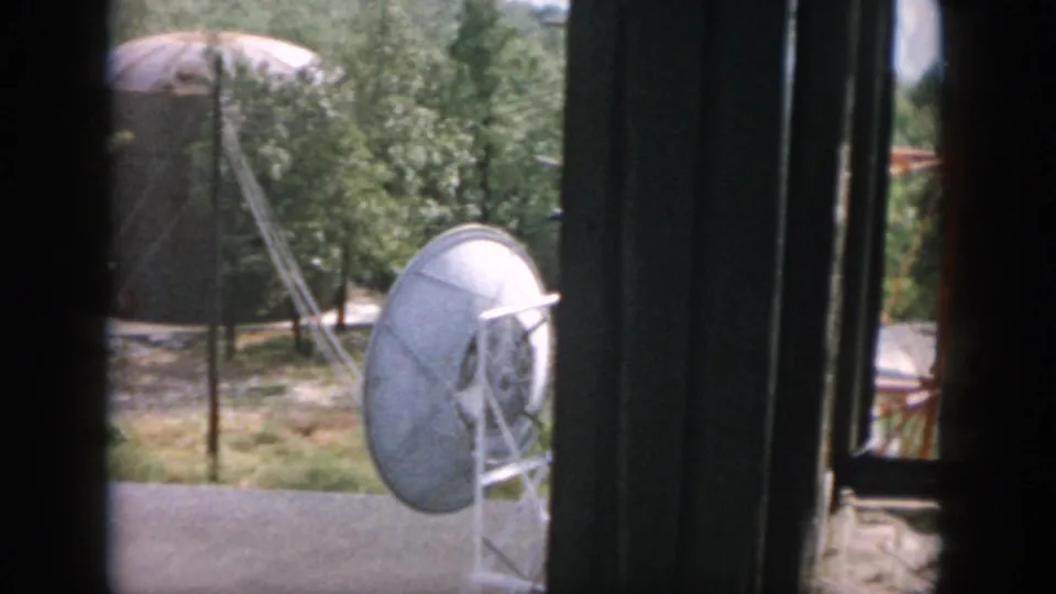 1958 Dayton 8mm frame showing a dish-like antenna, small water tower, trees, and a red structure beyond the window.
