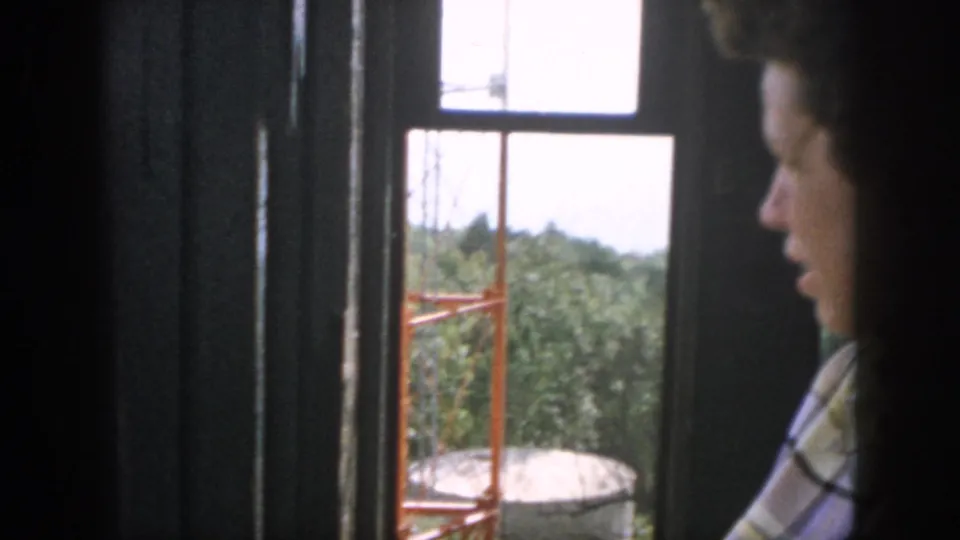 Young white man in a plaid shirt, filmed in profile inside a Dayton setting, looks out toward trees and industrial equipment in 1958 footage.