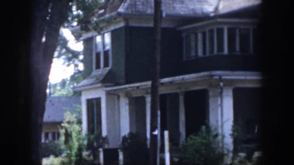 1958 Dayton, Ohio home-movie frame showing a two-story house with bay window, porch columns, dark siding, and trees in the foreground.