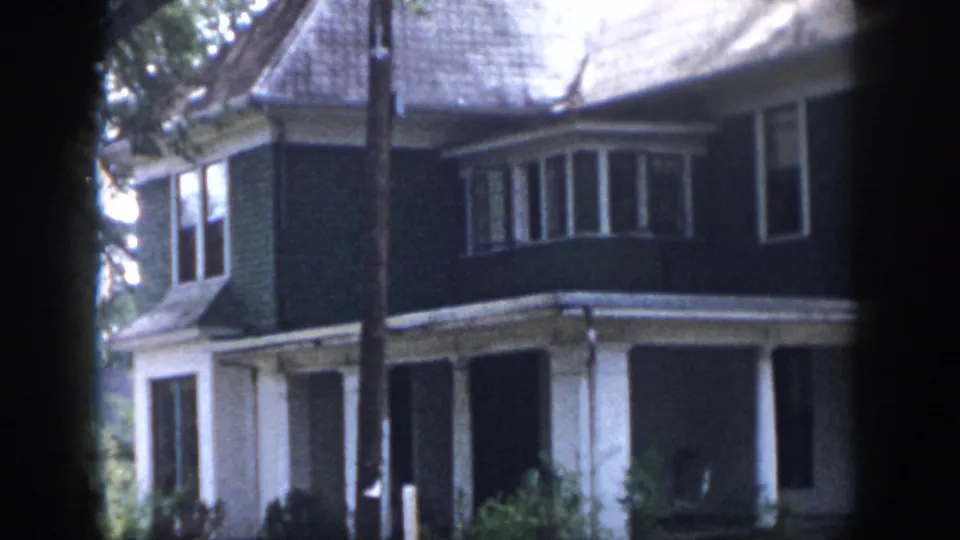 1958 Dayton, Ohio home-movie frame showing the house from a slightly shifted angle, with upper bay window, porch roof, tree trunk, and garden growth.