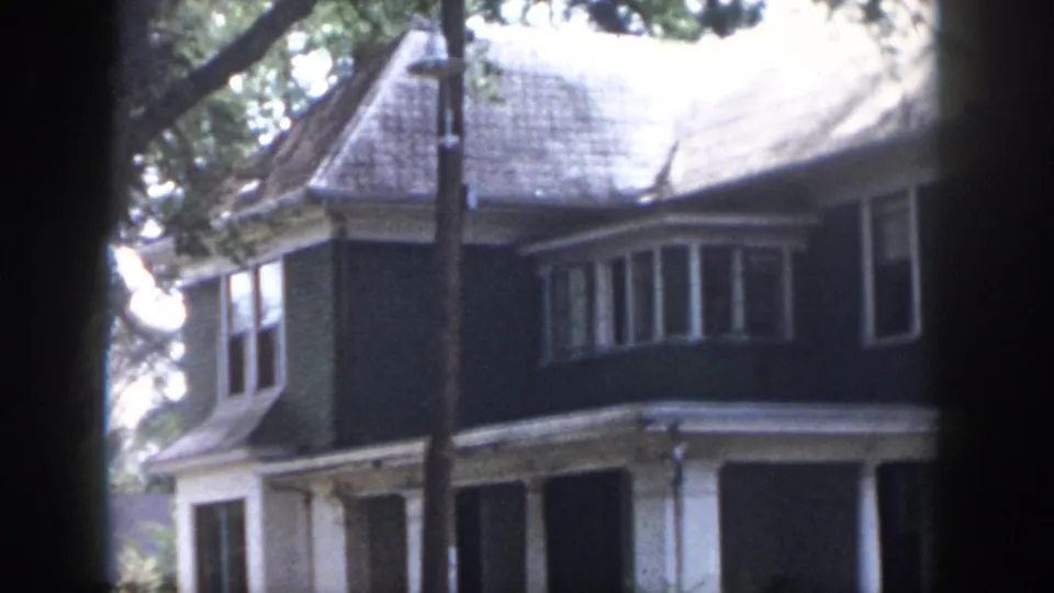 1958 Dayton, Ohio frame with soft focus and grain, showing a two-story house, bay window, porch columns, roofline, and tree branches in front.