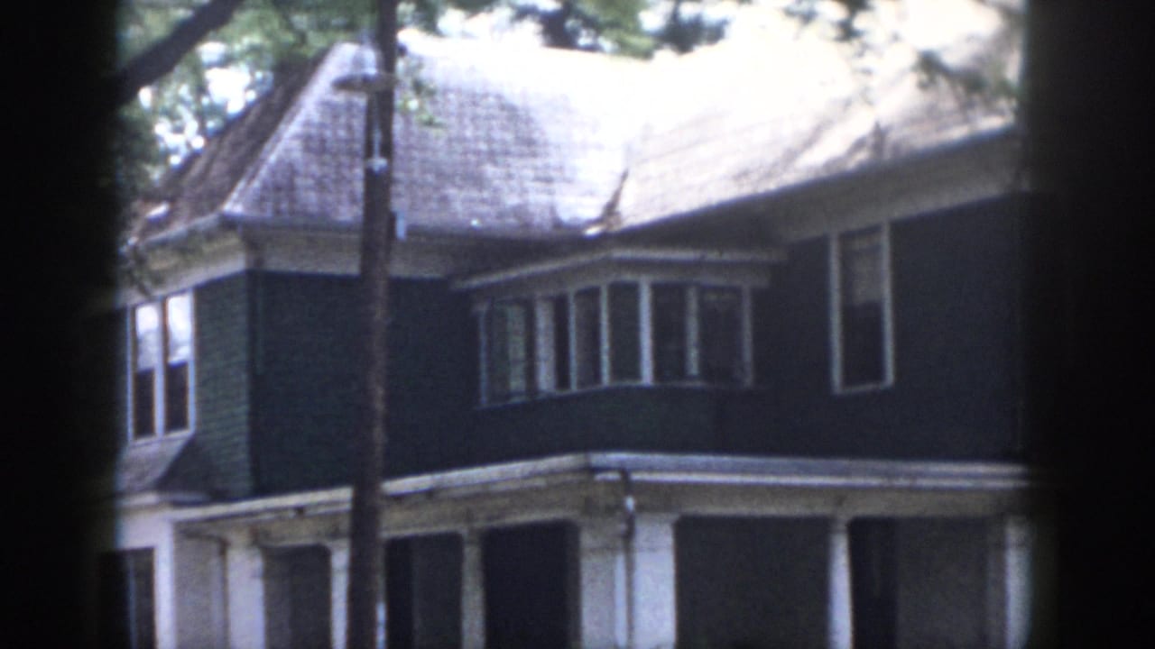 1958 Dayton home-movie frame showing a two-story house exterior with dark siding, white trim, a bay window, porch columns, and tree cover.