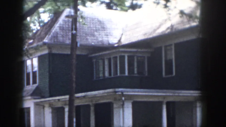 1958 Dayton home-movie frame showing a two-story house exterior with dark siding, white trim, a bay window, porch columns, and tree cover.