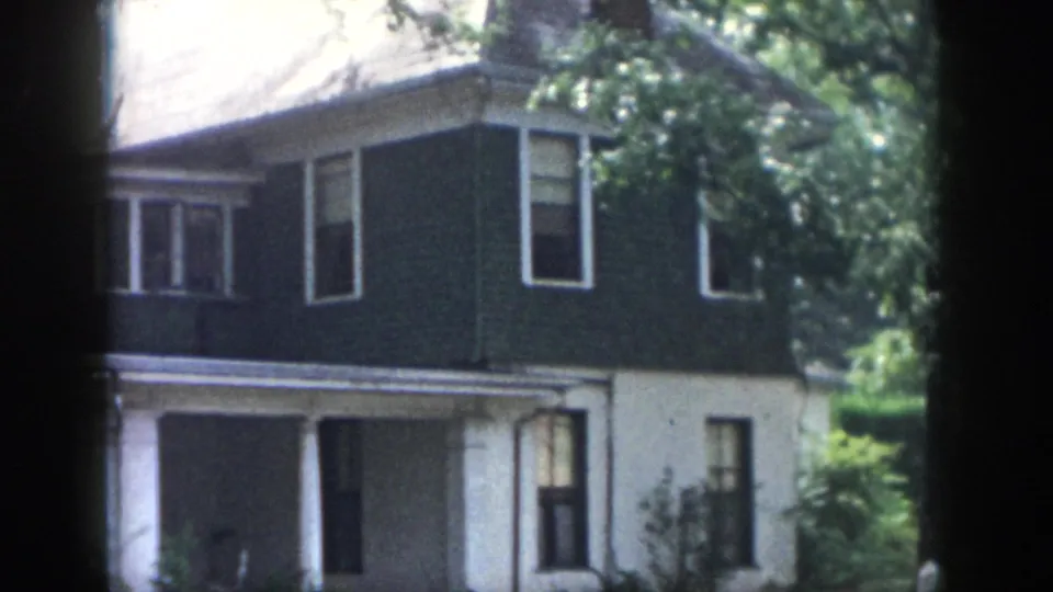 1958 Dayton frame showing the house from a slightly shifted angle, with side windows, porch roof, dark siding, white lower wall, and leafy yard.