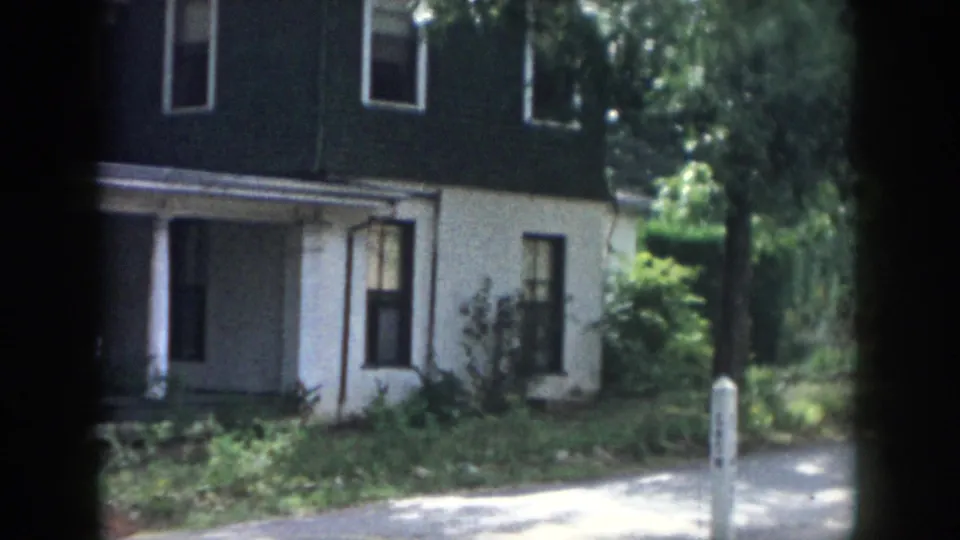 1958 Dayton home-movie frame showing the lower facade, porch entrance, dark upper siding, white lower wall, shrubs, trees, and a light driveway or walk.
