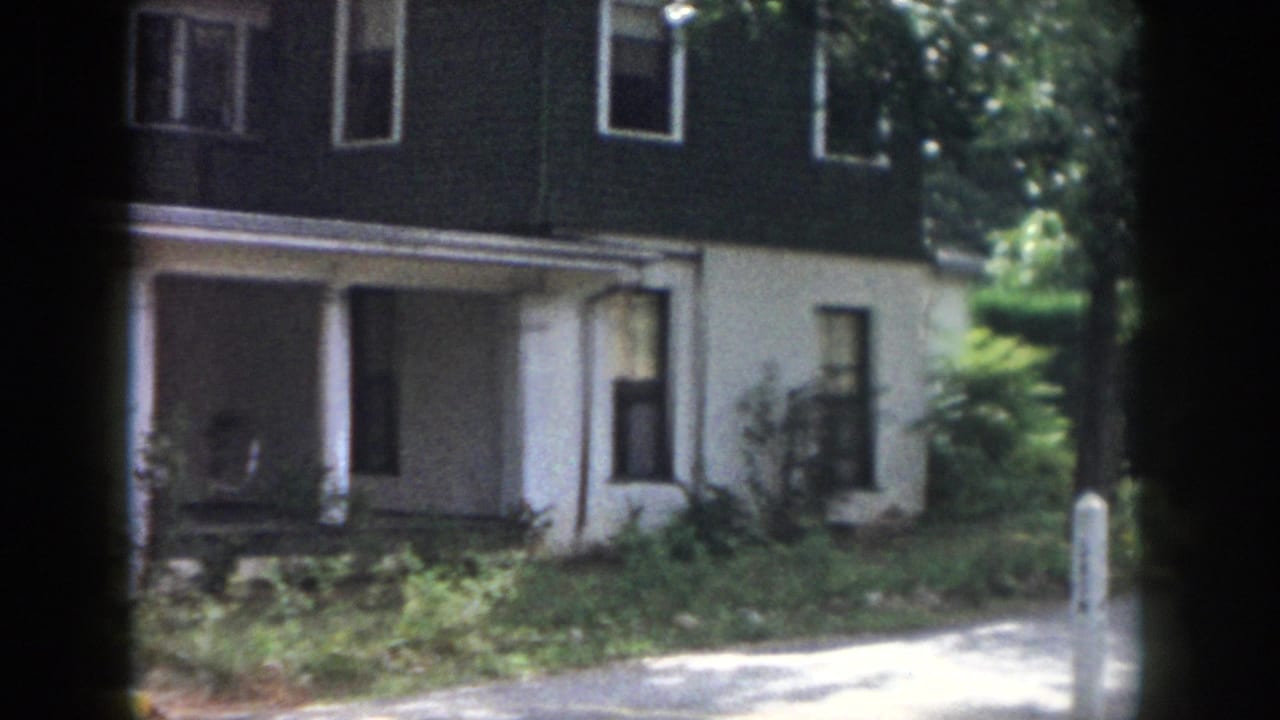 1958 Dayton home-movie frame showing a two-story dark-sided house with white trim, a covered porch, shrubs, and sidewalk shade.